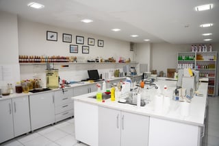 A modern cosmetics laboratory with white cabinets, central workbench, wall shelving displaying bottles and products, and framed certifications on the wall