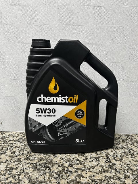 Black Chemist Oil 5W30 semi-synthetic motor oil bottle, 5 liters, on a speckled surface against white brick wall